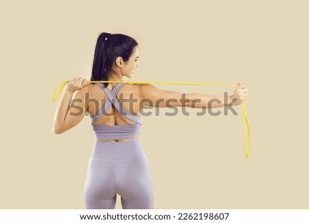 Happy attractive woman in modern sportswear enjoying regular fitness workout, training her body and doing physical exercises with elastic band. Backside view from behind isolated on beige background