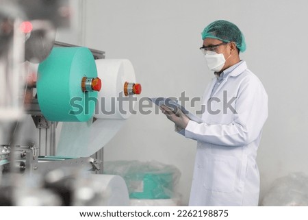 Worker man in personal protective equipment or PPE inspecting quality of mask and medical face mask production line in factory, manufacturing industry and factory concept.