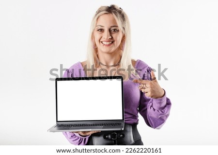 Blonde woman standing isolated over white background shows blank white mockup laptop screen to the camera and points with index finger and smiles at the camera. Saas or app advertisement.