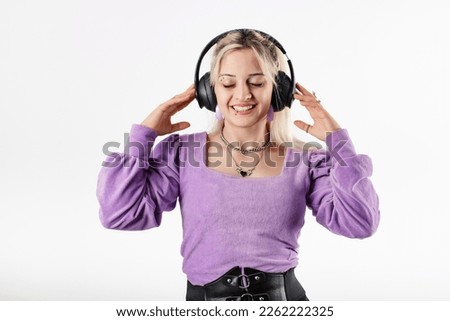 Young blonde girl smiling happy wearing lilac ribbed blouse isolated over white background closed eyes, listens to favorite music, touches headphones and enjoys it immensely.