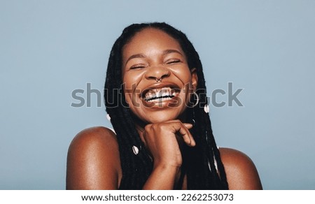 African woman with face piercings smiling cheerfully in a studio. Happy young woman wearing dreadlocks and piercing jewellery against a blue background. Female hipster feeling confident in her style. Royalty-Free Stock Photo #2262253073