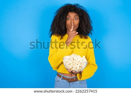 young woman with afro hairstyle wearing yellow sweater against blue background eating popcorn  makes silence gesture, keeps finger over lips. Silence and secret concept.