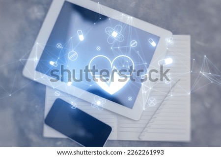 Top view of desktop with tablet, notepad and cellphone with glowing heart hologram on blurry background. Cardiology and doctor's office concept. Double exposure