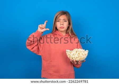 beautiful caucasian teen girl wearing pink sweater over blue wall purses lip and gestures with hand, shows something very little.