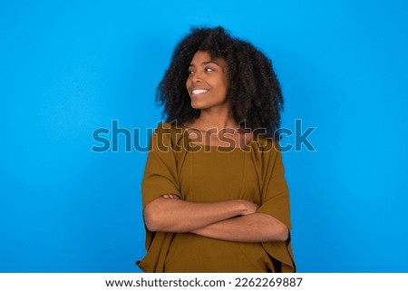 Dreamy rest relaxed young woman with afro hairstyle wearing brown shirt against blue background crossing arms, looks good copyspace Royalty-Free Stock Photo #2262269887