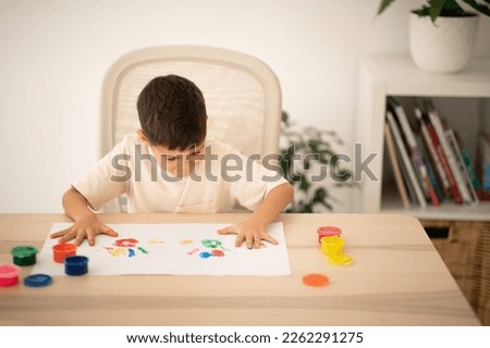 Glad european small kid draws picture with hands and paints at table in kindergarten, school or living room interior, empty space. Fun alone, fantasy at home, childhood and art, elementary education