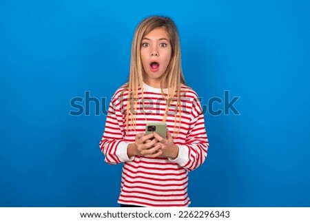 Shocked caucasian teen girl wearing striped t-shirt over blue wall opens mouth hold phone reading advert unbelievable big shopping prices