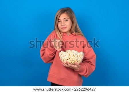 caucasian teen girl wearing pink sweater eating popcorn over blue wall makes bunny paws and looks with innocent expression plays with her little kid