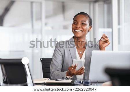 African black business woman using smartphone while working on laptop at office. Smiling mature african american businesswoman looking up while working on phone. Successful woman entrepreneur. Royalty-Free Stock Photo #2262320047