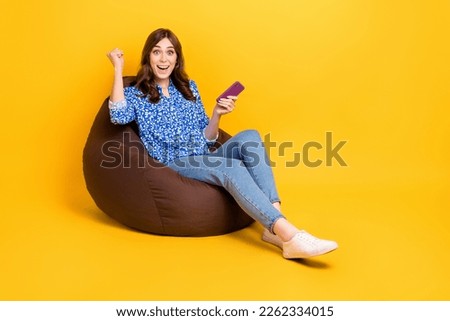 Full length photo of delighted crazy person sit comfort bag hold telephone raise fist success isolated on yellow color background