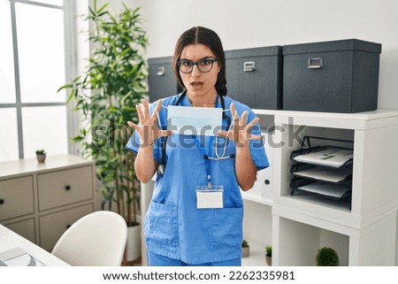 Young hispanic doctor woman holding safety mask at clinic in shock face, looking skeptical and sarcastic, surprised with open mouth 