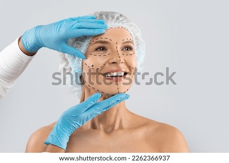 Cheerful excited middle aged woman in medical hat with pre surgery marks on her face looking at copy space for ad and smiling, surgeon hands in blue gloves touching female skin Royalty-Free Stock Photo #2262366937