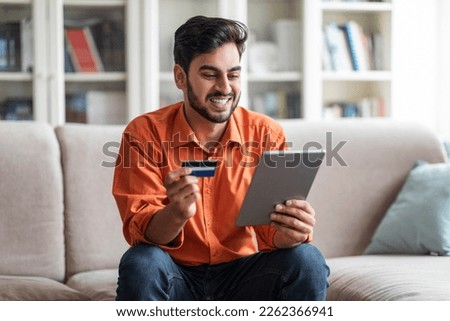 Cheerful smiling handsome young middle eastern man in smart casual sitting on couch in living room, using digital tablet and credit card, order food online, shopping on Internet from home, copy space Royalty-Free Stock Photo #2262366941
