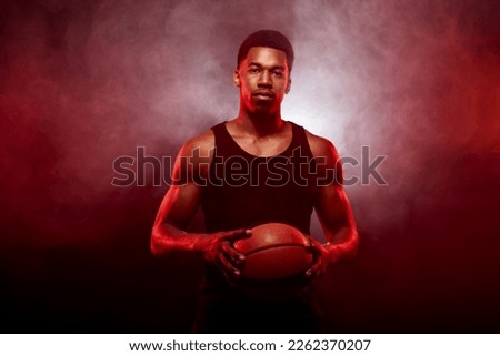 Basketball player side lit with red color holding a ball against smoke background. Serious concentrated african american man.
