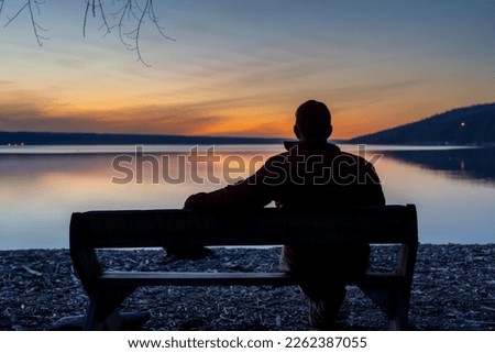Winter scene of a man with red jacket on a bench looking out over a lake at sunset.  Taking time for personal reflection, introspection, thinking about the past or the future.    Royalty-Free Stock Photo #2262387055