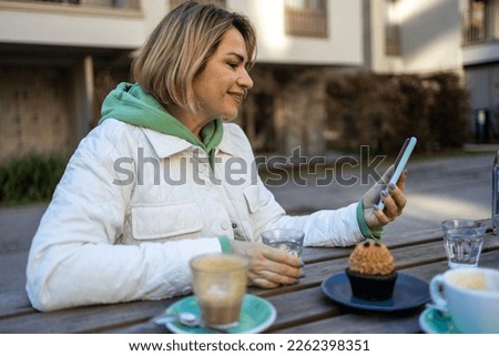 Blond middle aged woman having a video conference per smartphone while drinking a coffee in street caffe