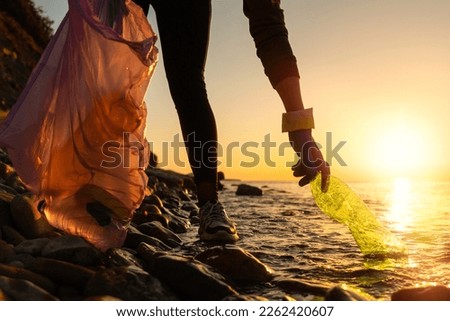 Pollution of ocean. A volunteer take a plastic bottle by the sea or river holding a plastic trash bag.  Low angle view. Coastal cleanup and garbage collection for recycling. World environment day 