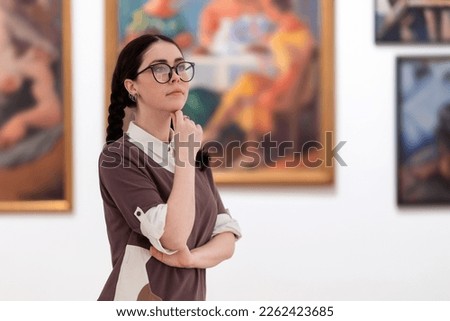 Cultural education and excursion. Portrait of caucasian pretty young woman wearing glasses contemplates arts. Defocused pictures in background. Concept of exhibition in gallery.