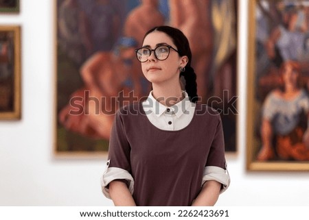 Cultural education. Portrait of caucasian pretty young woman student wearing glasses visiting excursion in museum, contemplates arts. Pictures in background. Concept of exhibition in gallery.