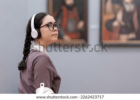 Side view of young Caucasian pretty woman wearing headphones, glasses and contemplates ancient artefacts. Student visiting arts exhibition. Concept of culture education and Museum day. Royalty-Free Stock Photo #2262423717