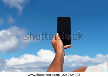Close-up of the arm of a tanned man taking a picture with his mobile phone of the blue sky with some clouds, during a sunny day in the touristic Canary Islands.