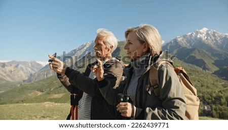 Senior caucasian couple having a nordic walk in mountains, then stopping to take a picture with smartphone. Old people travelling together after retirement - pension, tourism concept 