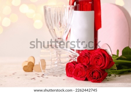 Bottle of wine, rose flowers, hearts and glasses on white table against blurred lights. Valentine's Day celebration