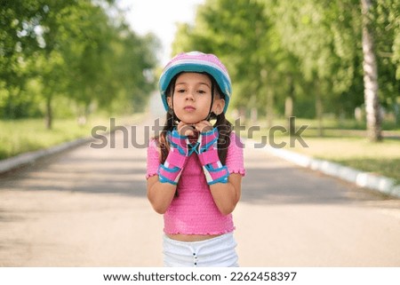 A little beautiful girl fastens a protective helmet. A child rides a skateboard in the park on a summer day Royalty-Free Stock Photo #2262458397