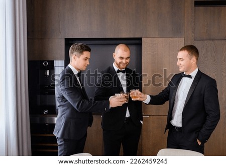 Front view of happiness groomsmen nd groom wearing in stylish black suits holding and clinking glasses, celebrating wedding day while standing on kitchen in apartment Royalty-Free Stock Photo #2262459245