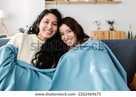 Two young female student roommates covered with blue blanket relaxing on sofa watching movie on television. Friendship and multimedia streaming entertainment concept Royalty-Free Stock Photo #2262469675