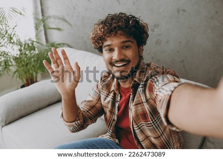 Close up happy young Indian man wears casual clothes do selfie shot pov mobile cell phone waving hand sits on grey sofa couch stay at home hotel flat rest relax spend free spare time in living room