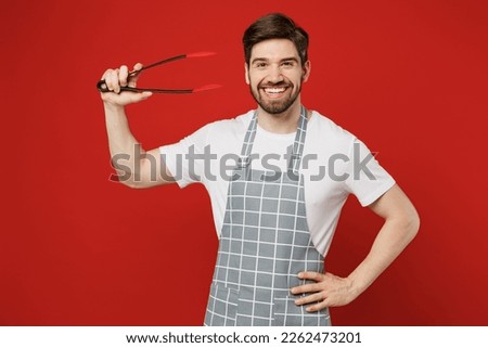 Young fun housewife housekeeper chef cook baker man wears grey apron hold in hand kitchen salad serving plastic pair of tongs for grill isolated on plain red background studio. Cooking food concept Royalty-Free Stock Photo #2262473201