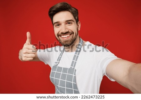 Close up fun young male housewife housekeeper chef cook baker man wear grey apron doing selfie shot pov on mobile cell phone show thumb up isolated on plain red color background. Cooking food concept