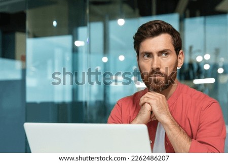 Close-up photo. A young male programmer, designer, freelancer in a red shirt who sits and works in the office at a laptop. He folded his hands together, looks thoughtfully and seriously to the side.