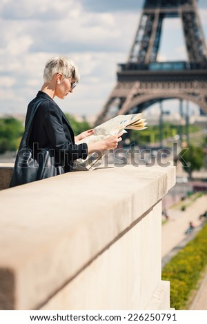 Young blonde woman looking at map of the city in front of the Eiffel Tower in Paris, France. Filtered image.