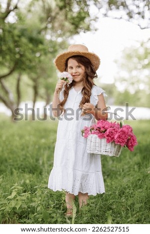 Portrait of smiling little girl wearing white lace dress and straw hat holds wicker basket with pink peonies flowers and walks outdoors in green park, enjoys the summer, warmth, flowers, freedom