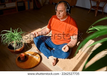 Mature middle-aged overweight man in wireless headphones relaxing at home with guided meditation, listening to relaxing music and meditating in lotus pose. Royalty-Free Stock Photo #2262527735