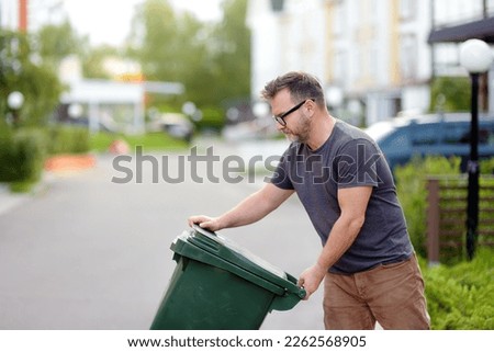 Mature man pulling out a large green plastic garbage container in front of the townhouse to the roadway of the street. Garbage collection and recycling trash, the zero waste concept. Waste managment Royalty-Free Stock Photo #2262568905