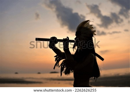 A silhouette of a Native American Indian plays music on a bamboo flute against a sunset background.