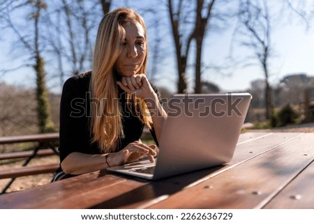 Concentrated blonde middle aged female freelancer sitting at wooden table and working on remote project on laptop in park