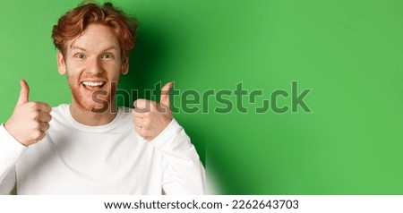 Emotions and fashion concept. Close up of cheerful redhead man showing thumbs-up and say yes, approve and like something cool, green background.