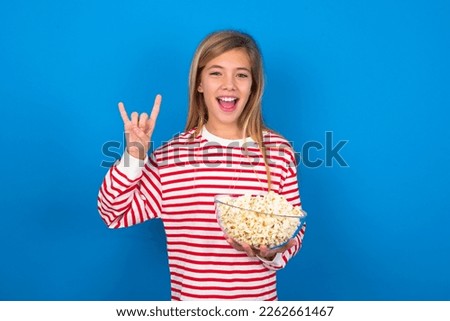 caucasian teen girl wearing striped t-shirt eating popcorn over blue wall doing a rock gesture and smiling to the camera. Ready to go to her favorite band concert. Royalty-Free Stock Photo #2262661467