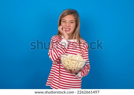Fearful caucasian teen girl wearing striped t-shirt eating popcorn over blue wall keeps hands near mouth, feels frightened and scared,  has a phobia,  Shock and frighted concept.