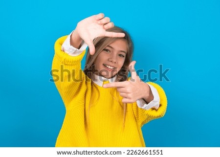 beautiful caucasian teen girl wearing yellow sweater over blue wall making finger frame with hands. Creativity and photography concept.