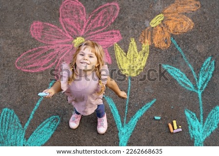 Little preschool girl painting with colorful chalks flowers on ground on backyard. Positive happy toddler child drawing and creating pictures on asphalt. Flower for mother's day