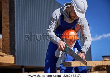 Father with toddler son building wooden frame house. Boy helping his daddy, using hand saw to cut boards on construction site, wearing helmet and blue overalls on sunny day. Carpentry, family concept. Royalty-Free Stock Photo #2262682171
