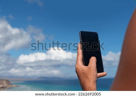 Close-up of a tanned man's arm looking at his mobile phone in the background of the white sandy beach and turquoise sea of Jandia, on a sunny day in the tourist resort of Fuerteventura, Canary Islands