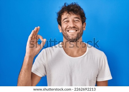 Hispanic young man standing over blue background smiling positive doing ok sign with hand and fingers. successful expression. 