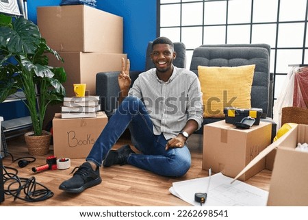 African american man sitting on the floor at new home smiling with happy face winking at the camera doing victory sign with fingers. number two. 