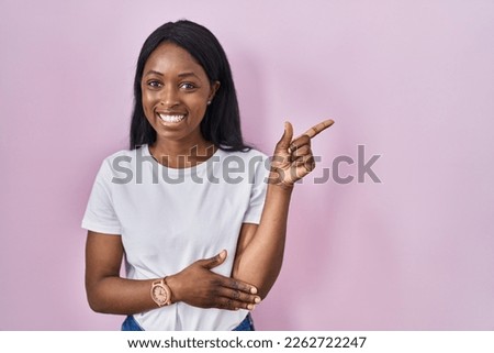 African young woman wearing casual white t shirt with a big smile on face, pointing with hand finger to the side looking at the camera. 
